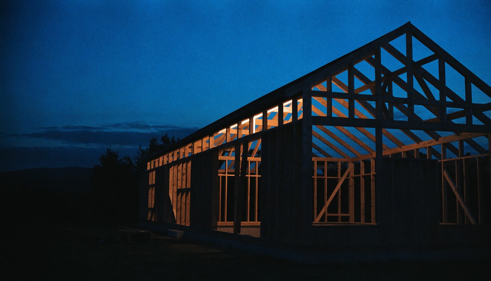 A timber-framed custom home under construction lit from within at dusk