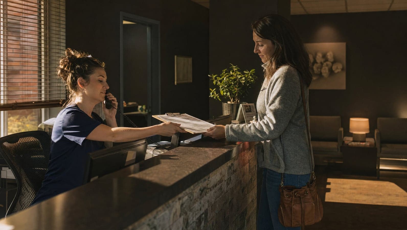 A dental receptionist answers the phone while handing a clipboard to a patient checking in at the front desk